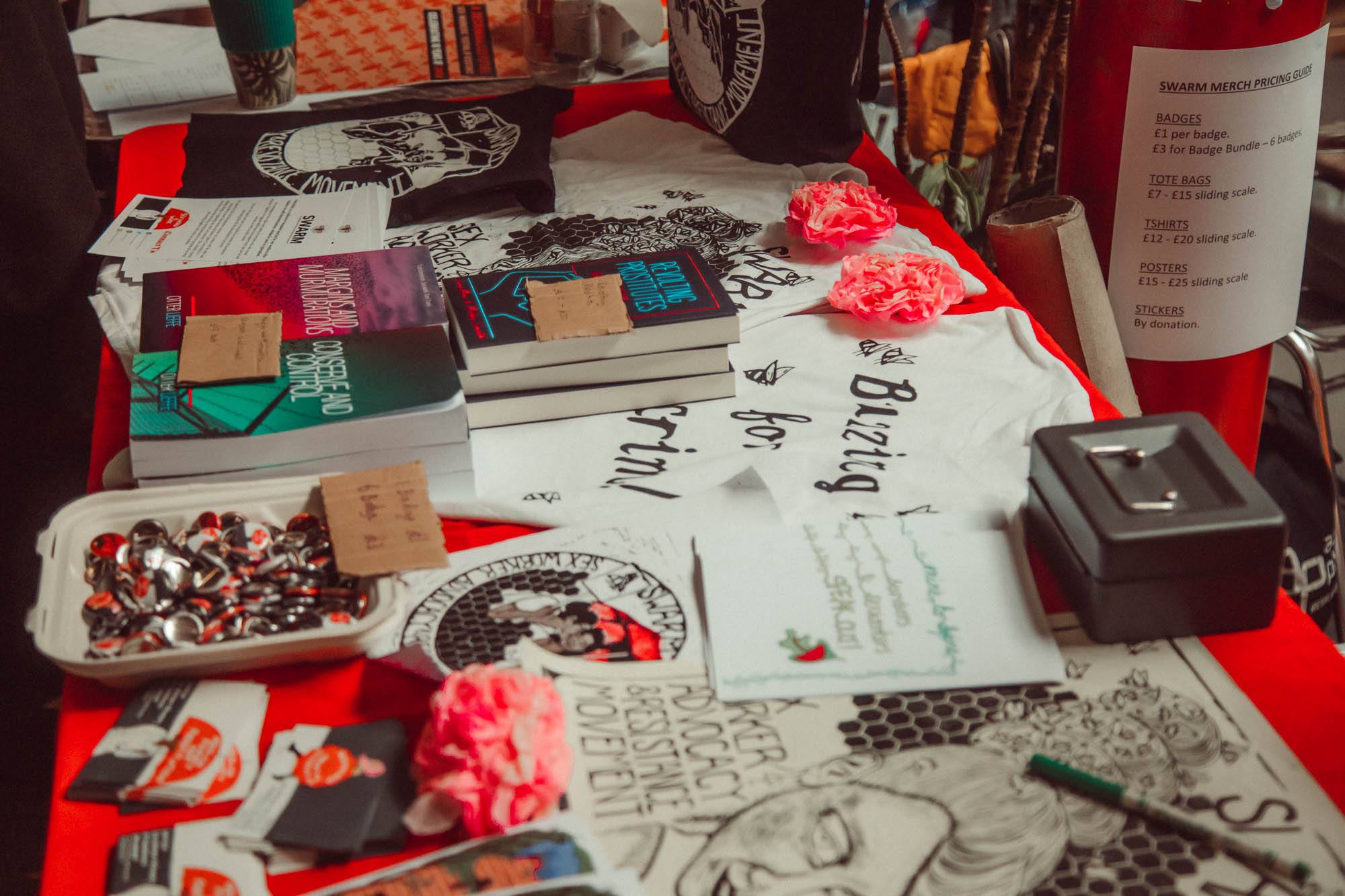A table featuring various merchandise from SWARM conference, including pints, badges, books and tshirts.