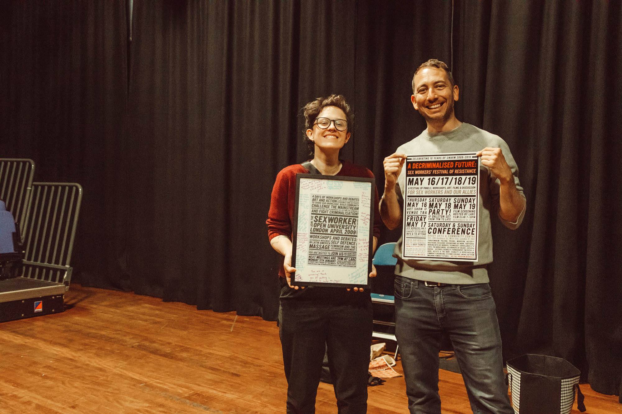 Two white people stand in front of a black cloth backdriop and wooden floors holding up SWARM conference posters. They are both smiling widely.