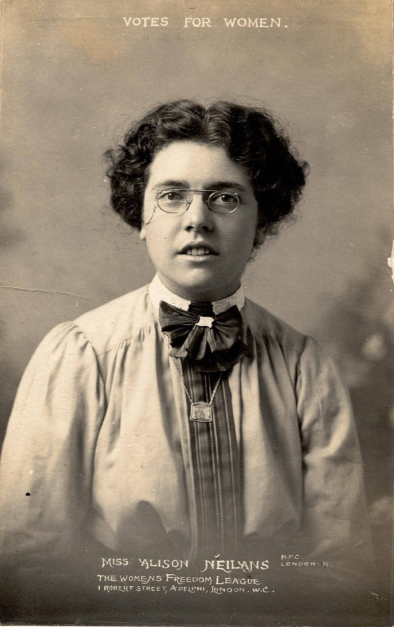 A black and white photograph of Alison Roberta Neilans, a white woman with wavy brown hair and glasses. She wears a loose-fitting, long sleeved blouse afixed with a bow at the collar.