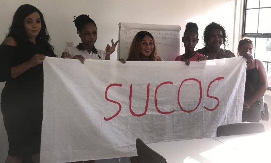 six women of colour stand in front of a table, next to a window, holding a large white banner that reads SUCOS in red writing.