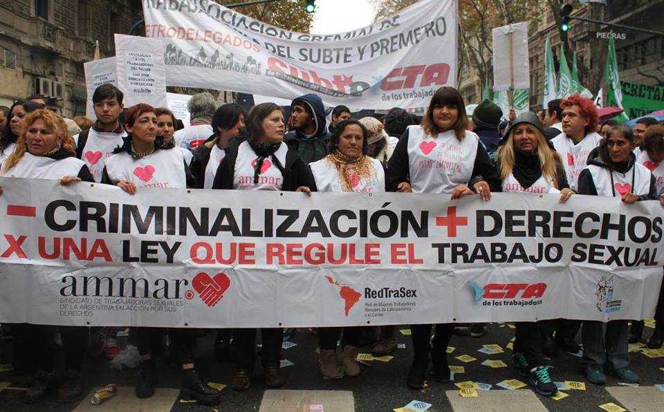 A large group of sex workers marching through the streets of Buenos Aires with a banner depicting the logos for AMMAR, RedTraSex and CTA (the Argentinian workers union).