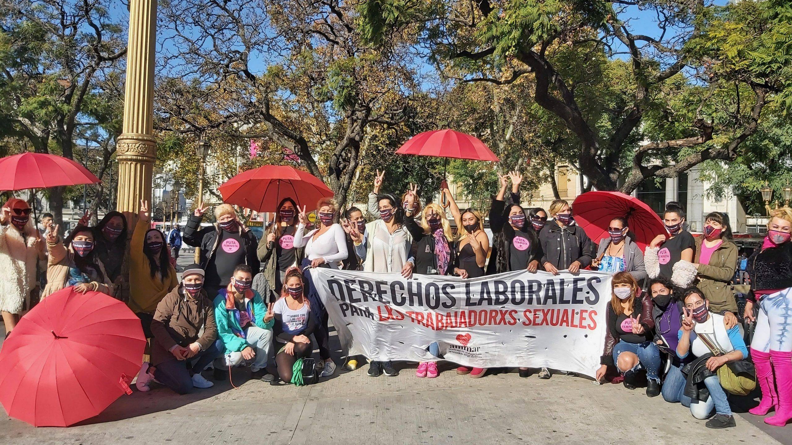 A group of about 25 people pose underneath some trees with red umbrellas and an AMMAR trade union banner that reads 