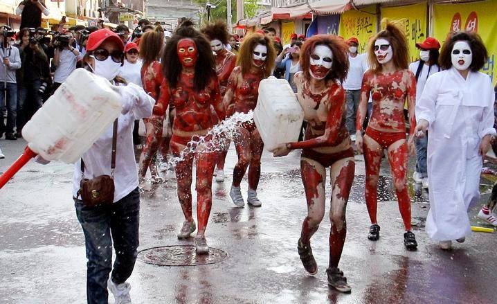 Eight women covered in red body paint and white face paint, with two of them in wearing white clothing, pour flammable liquid onto the street in front of onlookers.