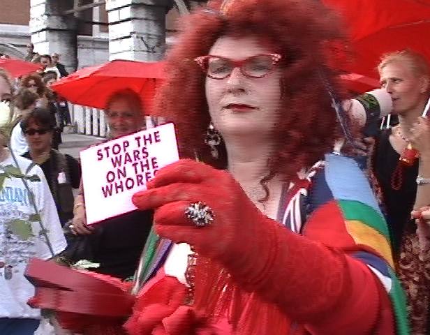 A woman with red hair and glasses is walking down the street. In her red gloved hand she holds a sign that says 'stop the wars on the whores'. People in the background are looking at her.