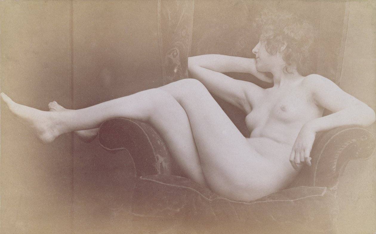 A sepia toned archival image of a young naked woman draped horizontally on a wingback sofa. She looks away from the camera.